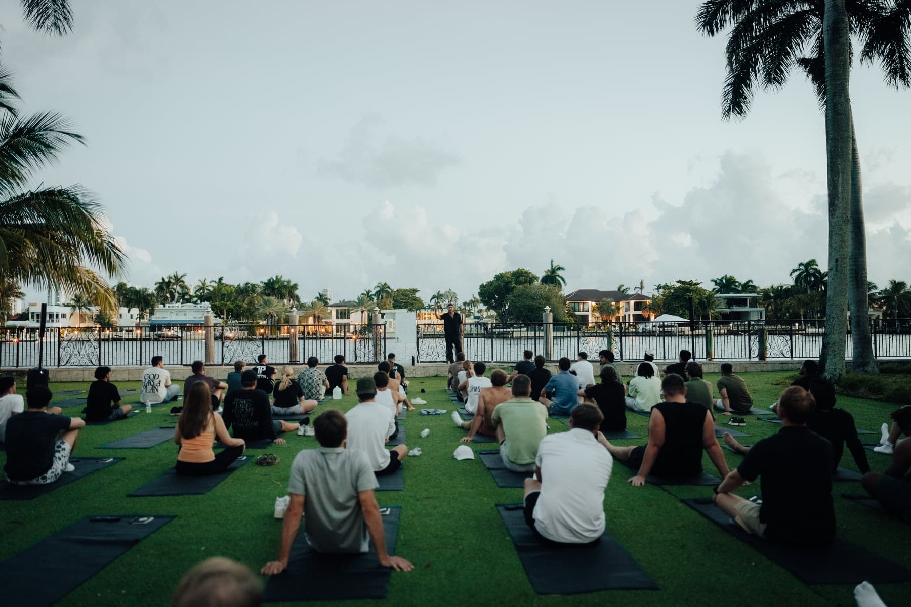 A group seated on mats across a waterfront lawn lined with palm trees