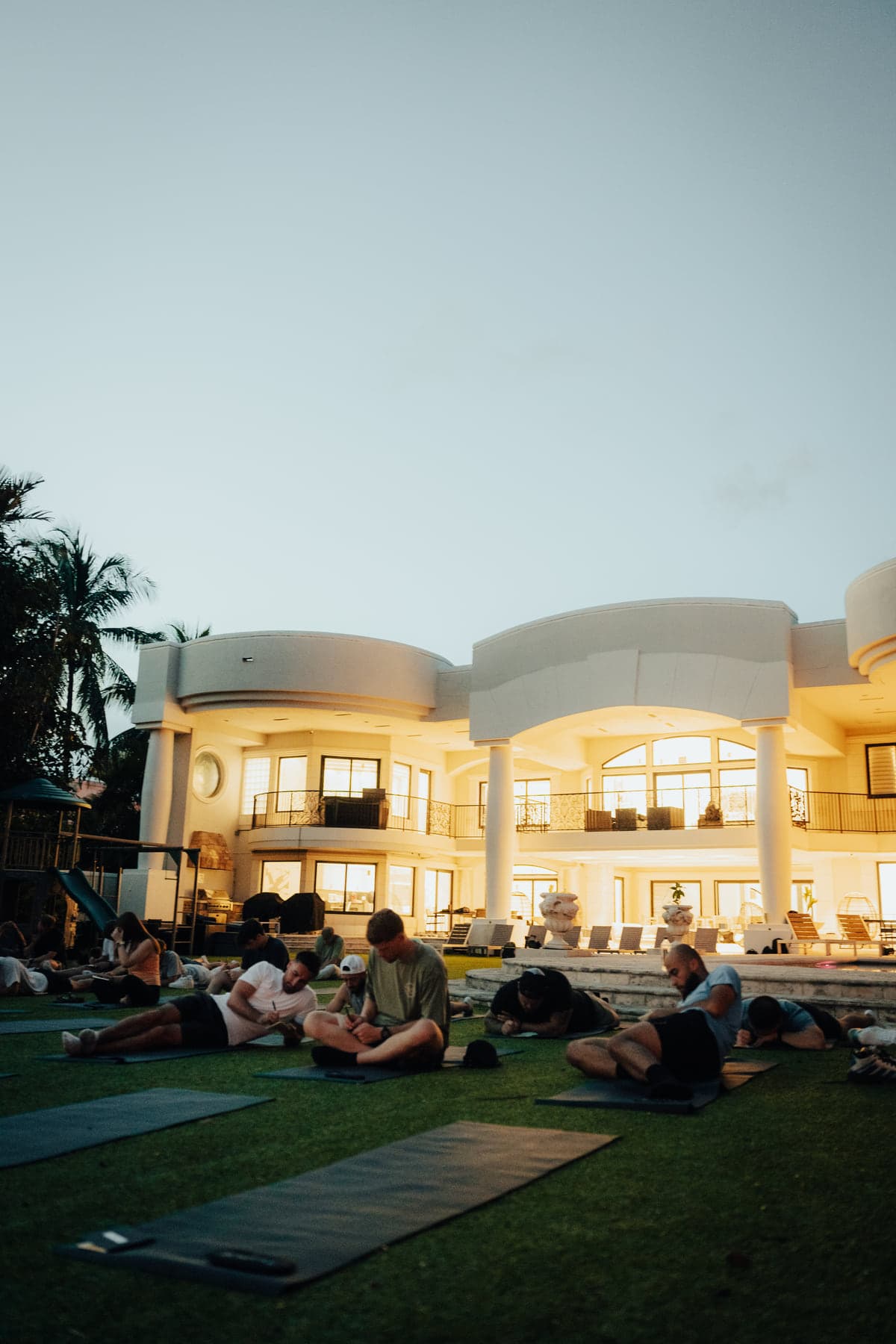 Participants resting on mats across an estate lawn as the house lights up at dusk