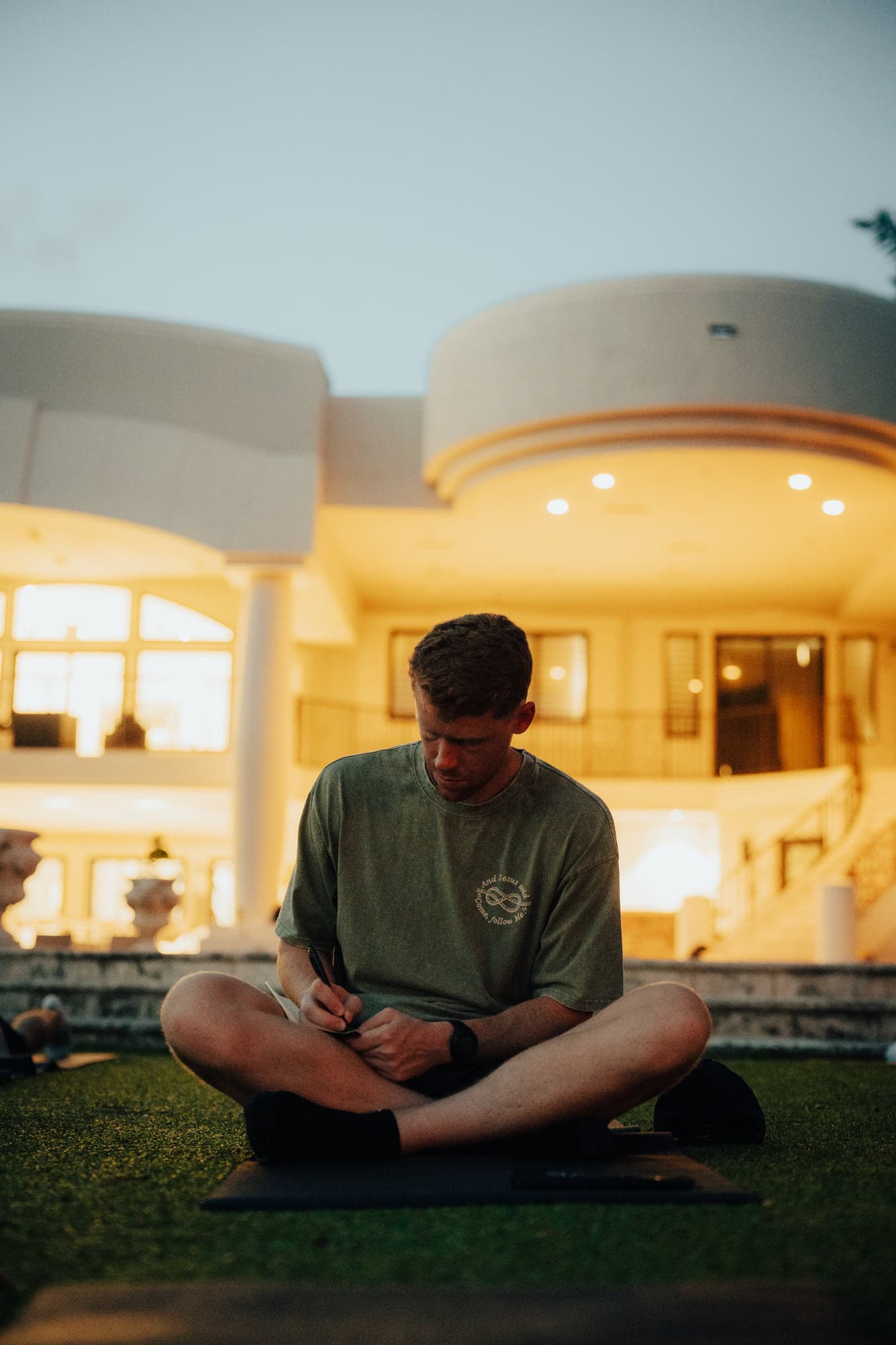 A participant journaling cross-legged on the lawn as the house behind him lights up at dusk