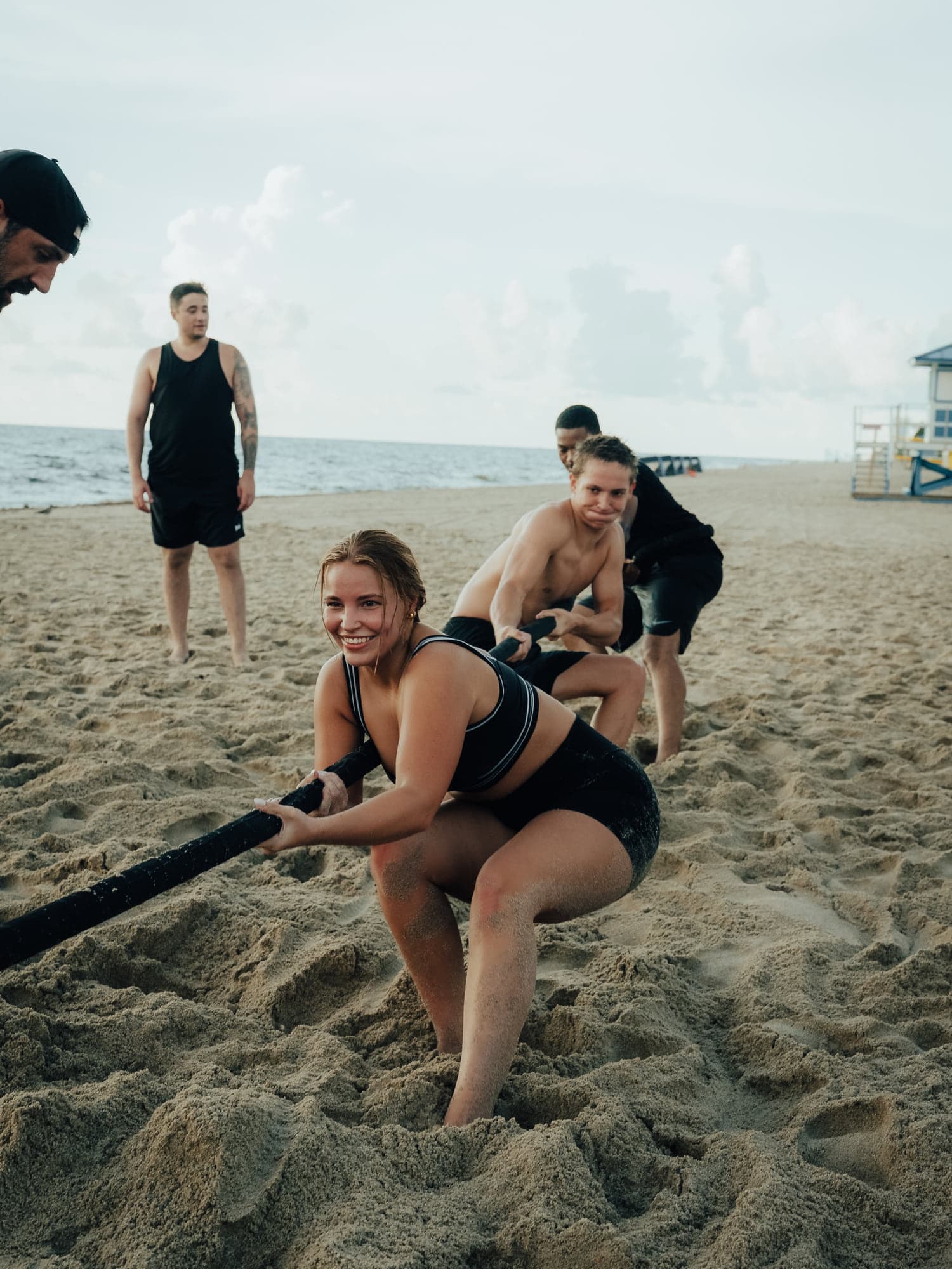 A participant leaning back on a heavy rope pull in the sand as teammates look on