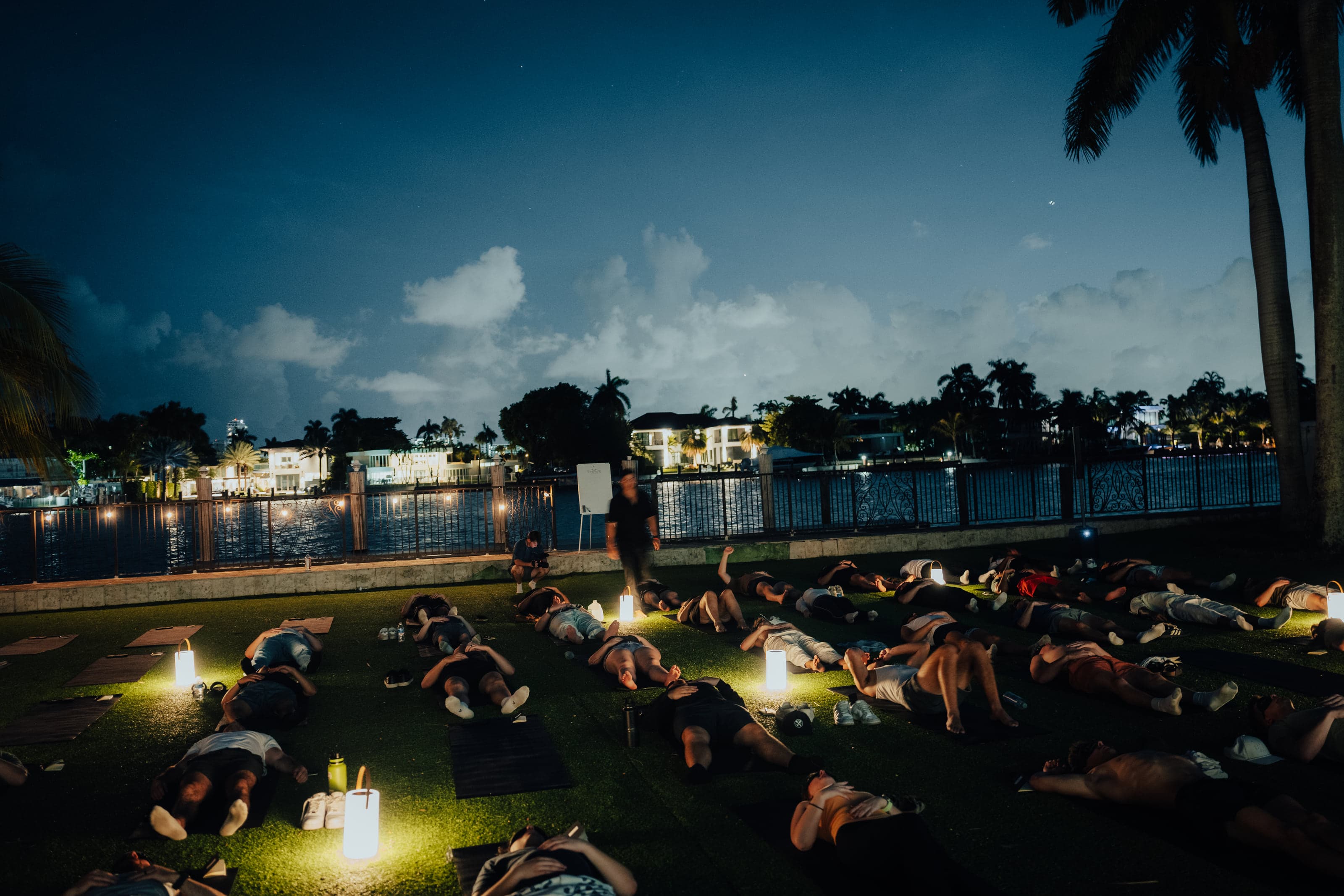 Participants lying on mats across a waterfront lawn at blue hour, lit by small lanterns, during a guided breathwork session