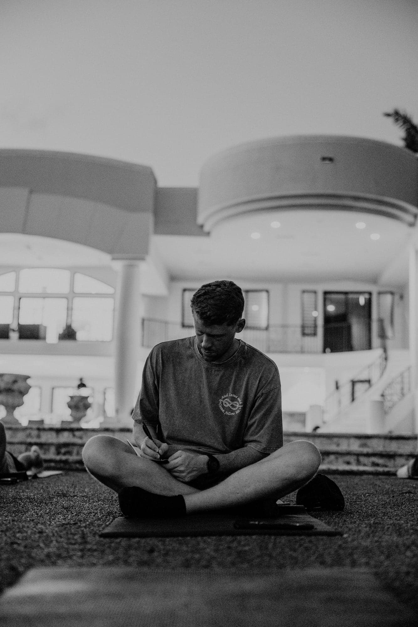 Black-and-white photograph of a participant sitting cross-legged on a lawn, writing in a journal in front of an illuminated house